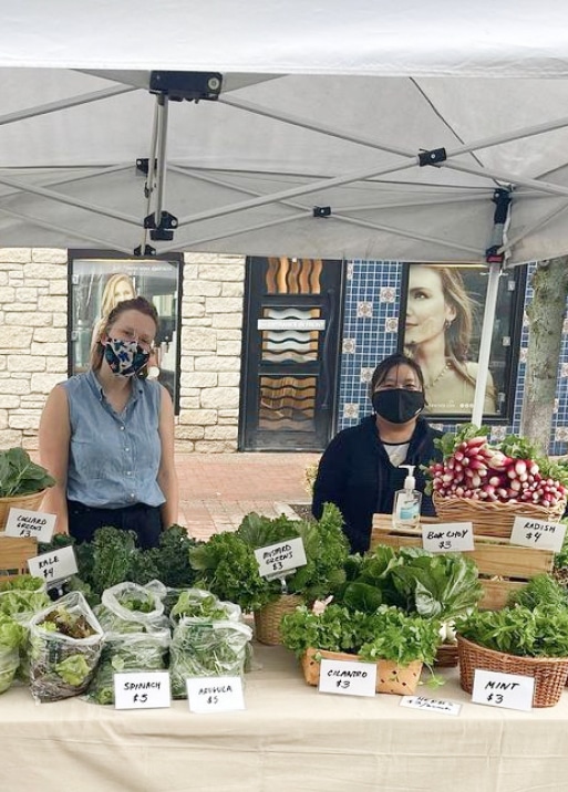 a woman wearing a face mask standing in front of a table full of vegetables.
